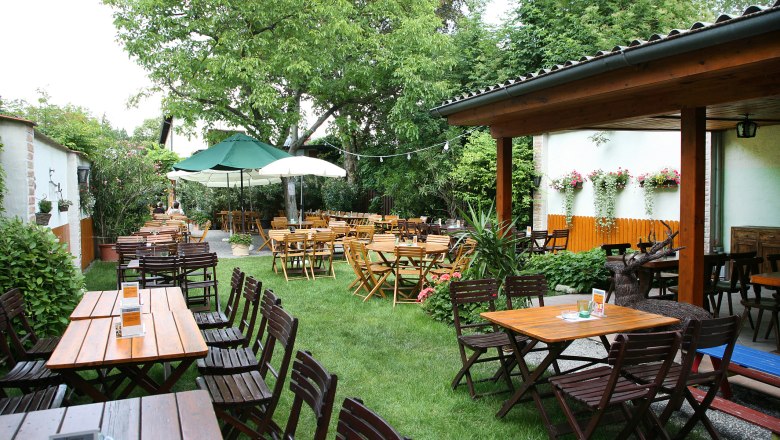 Garden, © Zierer A cozy beer garden with wooden tables and chairs, surrounded by green trees and plants.
