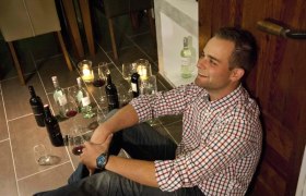 Der Winzer Andreas, © Weingut Artner Ein Mann sitzt lächelnd auf dem Boden, umgeben von Weinflaschen und Gläsern.