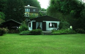 Haus, © Haus Ryba Ein kleines Haus mit Garten und Terrasse, umgeben von Bäumen und einem größeren Gebäude im Hintergrund.