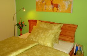 Die kleine Pension, © Die kleine Pension Gemütliches Schlafzimmer mit grünem Bettzeug, Holzkopfteil, grüner Wand und Hirschbild. Eine Lampe und Blumen stehen auf dem Nachttisch.