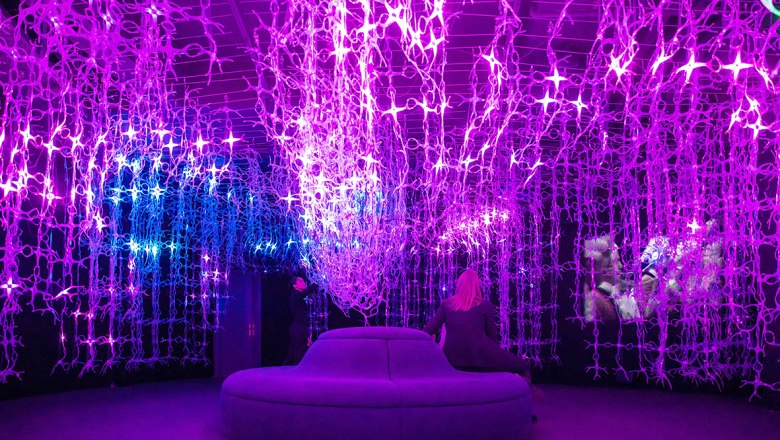 Mythos Mozart, © Mythos Mozart Ein Raum mit violetten und blauen Lichterketten, die von der Decke hängen. Eine Person sitzt auf einem runden Sofa.