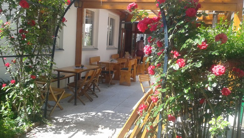 Rose arch, © Dominik Herzog A rose arch with red roses leads to an outdoor area with wooden tables and chairs.