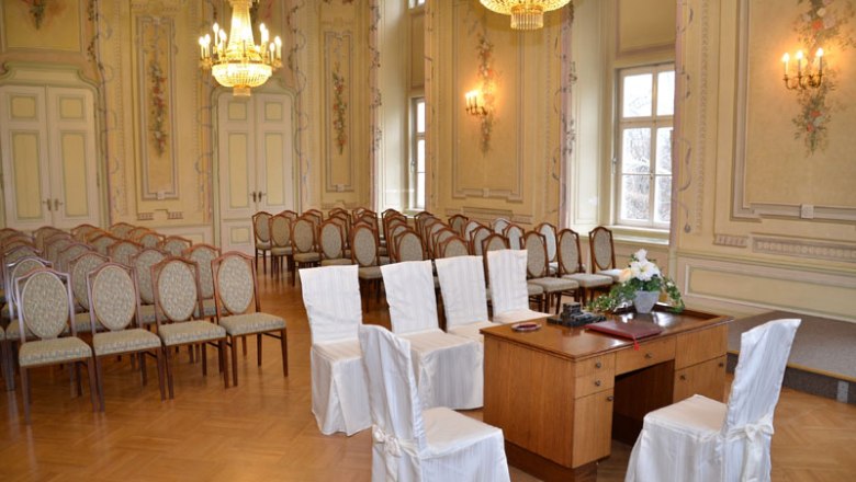 Ballroom with covers, © Silke Ebster Elegant ballroom with chairs covered in white covers and a wooden table. Chandeliers and ornate walls adorn the room.
