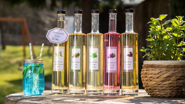 Selbstgemachte Sirupe aus dem Kräutergarten vom Gasthaus Rath-Smetana, © Niederösterreich Werbung/Kurt Pinter Fünf Flaschen mit selbstgemachtem Sirup und ein blaues Glas mit Strohhalmen auf einem Holztisch im Freien.