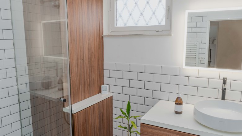 Bathroom, © Theodor Haberhauer Modern bathroom with wooden cabinets, white tiled wall, round washbasin and illuminated mirror.