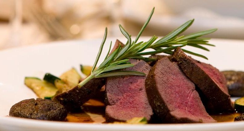 Gourmet-Menü, © F. Bläuel GesmbH Ein Gourmet-Gericht mit rosa gebratenem Fleisch, garniert mit Rosmarin, auf einem Teller mit Zucchini und Pilzen.