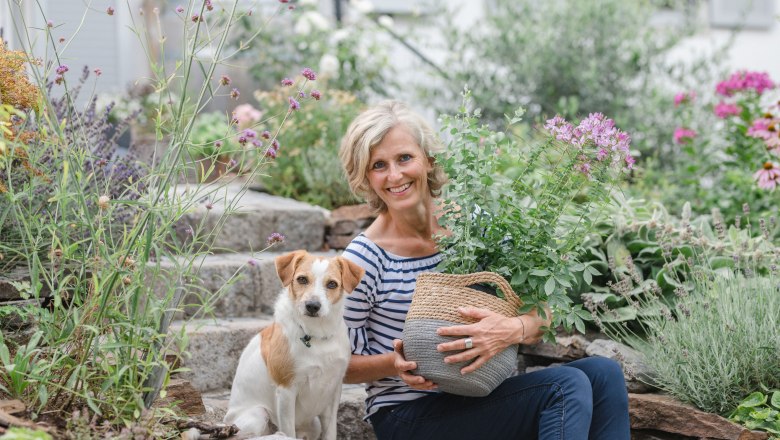 Sommergarten Christines Paradies, © Eva Lekey Fotografie Eine Frau sitzt mit einem Hund in einem blühenden Garten auf Steinstufen.