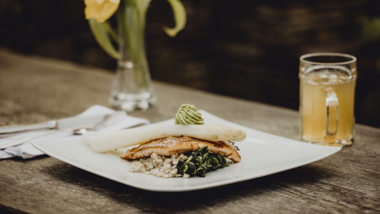 Zarter Fisch trifft knackigen Spargel, © Niederösterreich Werbung/Sophie Menegaldo Gourmetgericht mit Spargel, Fisch und Gemüse auf Holztisch, gelbe Blume im Hintergrund.
