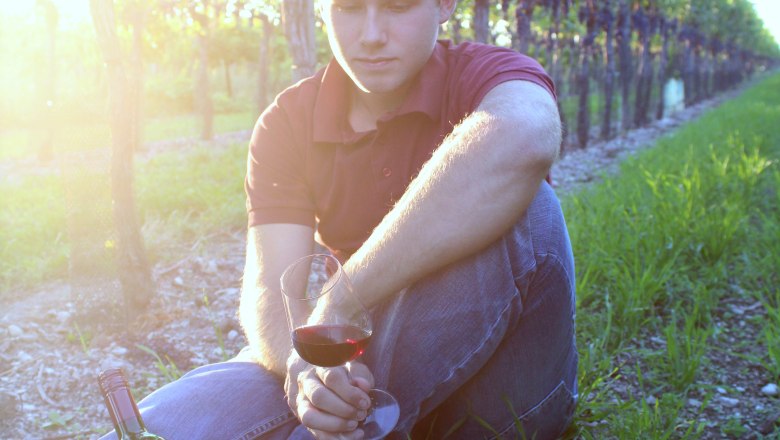 The junior boss, © A.Knötzl A young man is sitting in a vineyard holding a glass of red wine. Next to him is a bottle of wine. The sun is shining through the leaves.