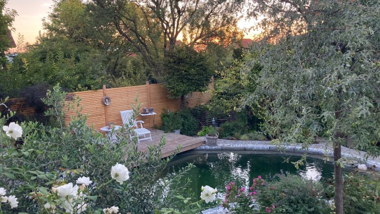 Christines Paradies, © Christine Hammerschick Ein idyllischer Garten mit Teich, Holzterrasse und weißen Gartenstühlen, umgeben von üppiger Vegetation und blühenden Blumen.