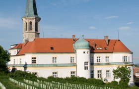 Das Schloss Gumpoldskirchen, © Schloss Gumpoldskirchen Betriebs-GmbH Schloss Gumpoldskirchen mit rotem Dach und Turm, umgeben von Weinbergen unter blauem Himmel.