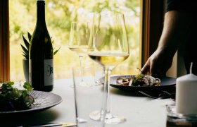 Restaurant im Weingut Johnneshof Reinisch, © Julius Hirtzberger Ein gedeckter Tisch mit Weinflasche, Weingläsern und einem Teller mit Essen in einem Restaurant.