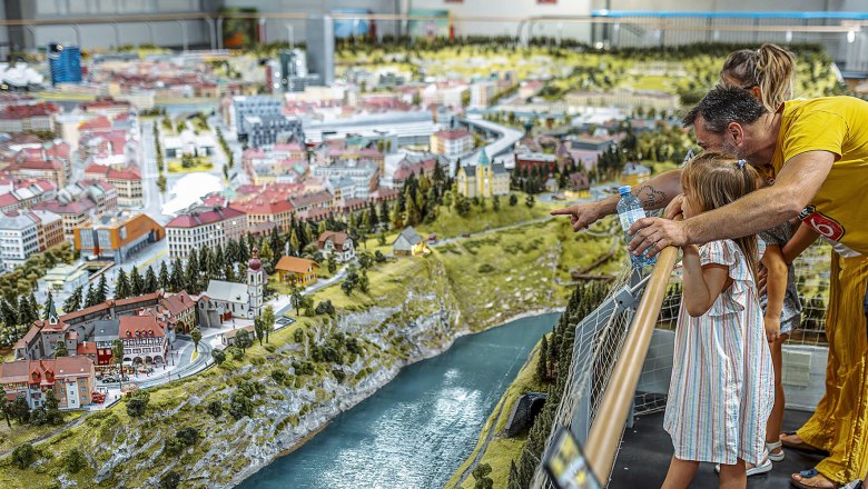 Königreich der Eisenbahnen, © © Königreich der Eisenbahnen, Adam & Costey studio Familie betrachtet Modellbahnanlage mit Stadt und Fluss.