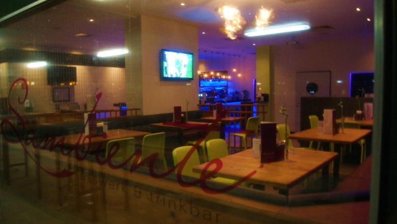 Lokal, © sambiente Innenansicht eines leeren Restaurants mit Holztischen und bunten Stühlen, Fernseher an der Wand und beleuchteter Bar im Hintergrund.