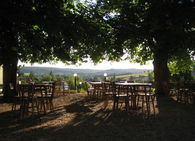 Garten, © Meierei Gaaden Ein schattiger Garten mit Holztischen und -stühlen unter großen Bäumen, mit Blick auf eine hügelige Landschaft im Hintergrund.