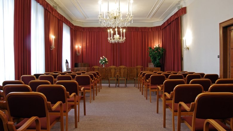 Wedding hall registry office Klosterneuburg, © Benjamin Zibuschka An elegant wedding hall with red curtains, chandeliers and rows of chairs.