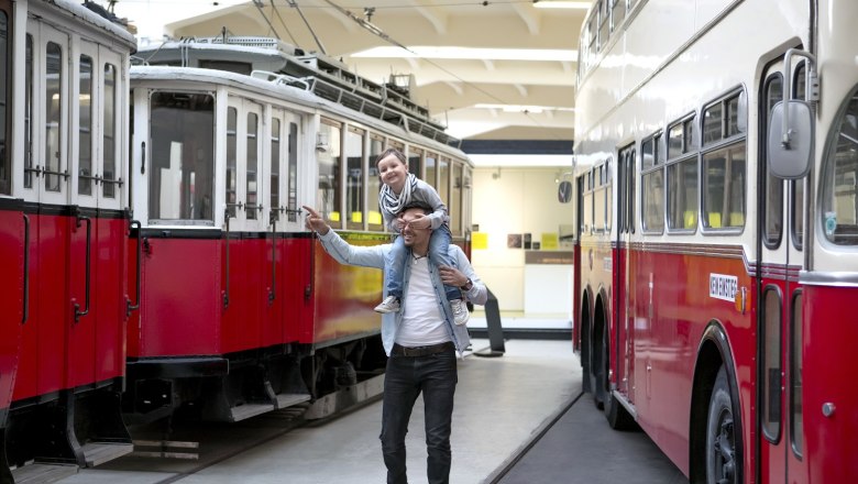 Verkehrsmuseum der Wiener Linien, © Wiener Linien Ein Mann trägt ein Kind auf den Schultern im Verkehrsmuseum der Wiener Linien, umgeben von historischen Straßenbahnen und Bussen.
