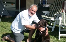 Tierfreund, © L.G Ein Mann kniet neben einem braunen Hund auf einer Wiese vor einem Wohnwagen.