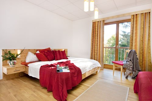 Room, © Brandtner Bright bedroom with double bed, red pillows and blanket, wooden floor and large window with curtains.