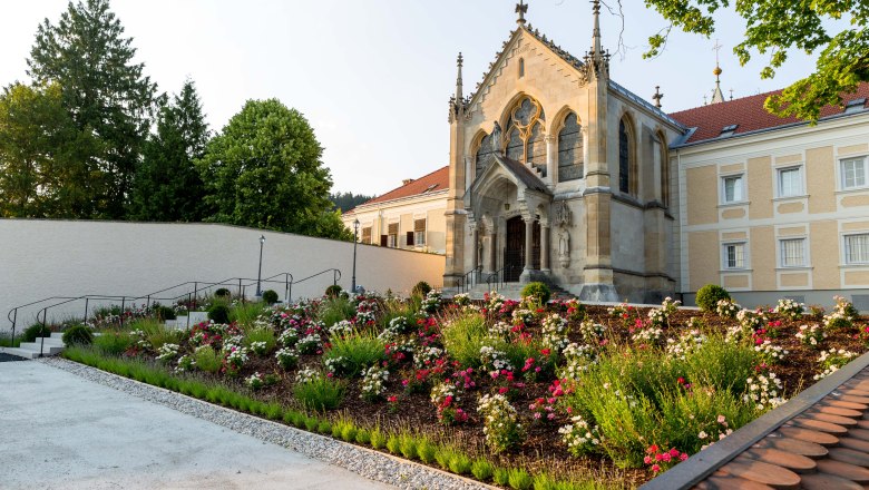 KirchemitGartenanlage, © SusanneHammerle Eine Kirche mit gotischen Elementen und einem gepflegten Garten voller Blumen im Vordergrund.
