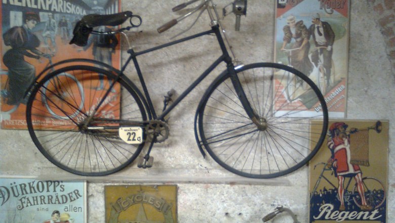 Fahrradmuseum Schloss Vösendorf, © Fahrradmuseum Schloss Vösendorf Altes Fahrrad an einer Wand mit Vintage-Werbeplakaten im Fahrradmuseum Schloss Vösendorf.