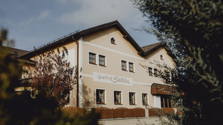 Wirtshaus mit großem Spielplatz, © Niederösterreich Werbung/Sophie Menegaldo Ein traditionelles Gasthaus mit der Aufschrift 'Gasthof Skilitz', umgeben von Bäumen und einem Holzzaun.