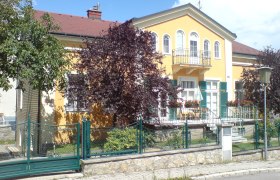 Hawlik, © Ferienwohnungen Hawlik Gelbes Haus mit Balkon und grünem Zaun, umgeben von Bäumen.