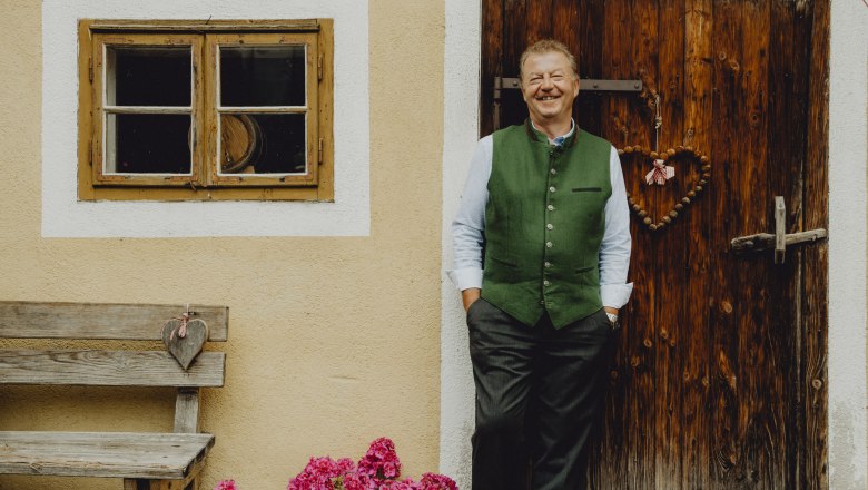 Landlord Anton Schilling, © Niederösterreich Werbung/Rita Newman A man in traditional dress stands smiling in front of a wooden door next to a window and a bench with flowers.