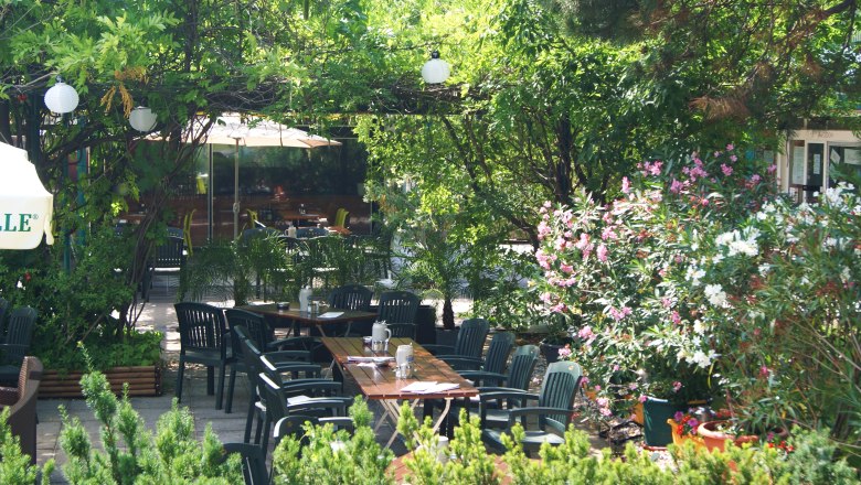 Garten, © sambiente Ein grüner Garten mit Tischen und Stühlen, umgeben von Bäumen und blühenden Sträuchern.