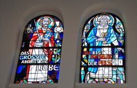 Windows, © Pfarre St. Jakobus Two colorful stained glass windows with religious figures and inscriptions.
