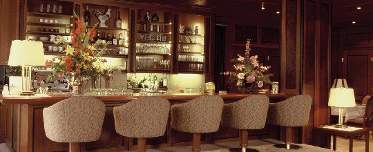 Bar, © Hotel Jagdhof Elegante Bar mit Holzverkleidung, Blumenarrangements und gepolsterten Barhockern.