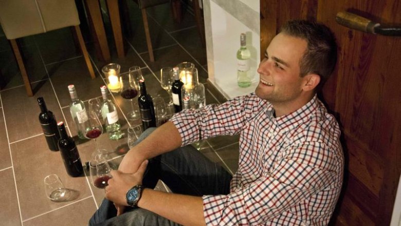 Der Winzer Andreas, © Weingut Artner Ein Mann sitzt lächelnd auf dem Boden, umgeben von Weinflaschen und Gläsern.