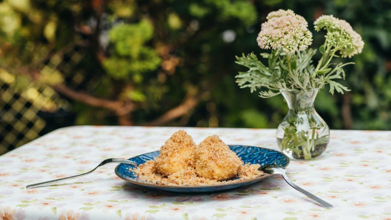 Plum dumplings, © Niederösterreich Werbung/Daniela Führer A plate with two plum dumplings on a table with a floral patterned tablecloth, next to it a vase with flowers.