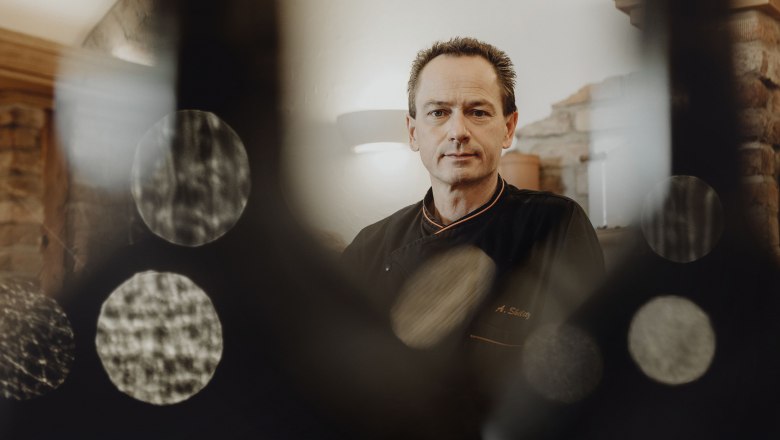 Wirt Alfred Skilitz, © Niederösterreich Werbung/Sophie Menegaldo Ein Mann in schwarzer Kochkleidung steht in einem rustikalen Raum, umgeben von unscharfen Lichteffekten.