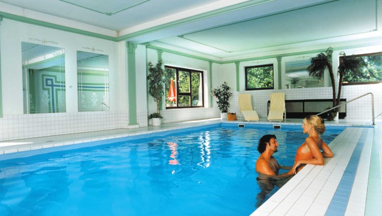 Pool, © Das Steinberger Ein Paar im Pool eines eleganten Innenbereichs mit Pflanzen und Liegestühlen.