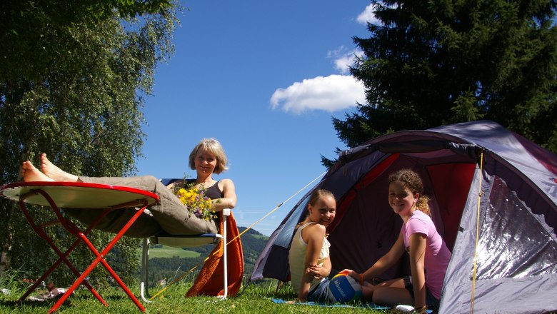 Camping life, © Reinhard Wallentin A woman relaxes on a camping chair next to a tent, while two children sit in the tent. Trees and blue sky can be seen in the background.