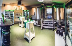 Innenansicht einer kleinen Brauerei mit Edelstahlkesseln und Kühlschränken voller Getränke.