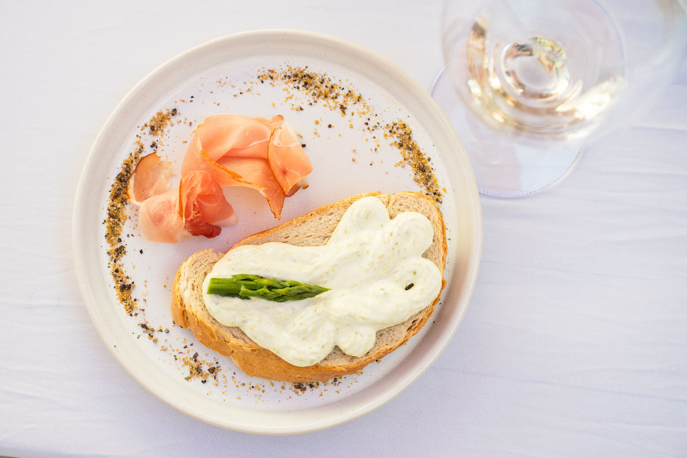 Weiß gedeckter Tisch mit weißem Teller in freundlicher Atmosphäre; darauf Baguette mit heller Creme, grünem Spargel und Prosciutto, daneben ein Glas Weißwein.