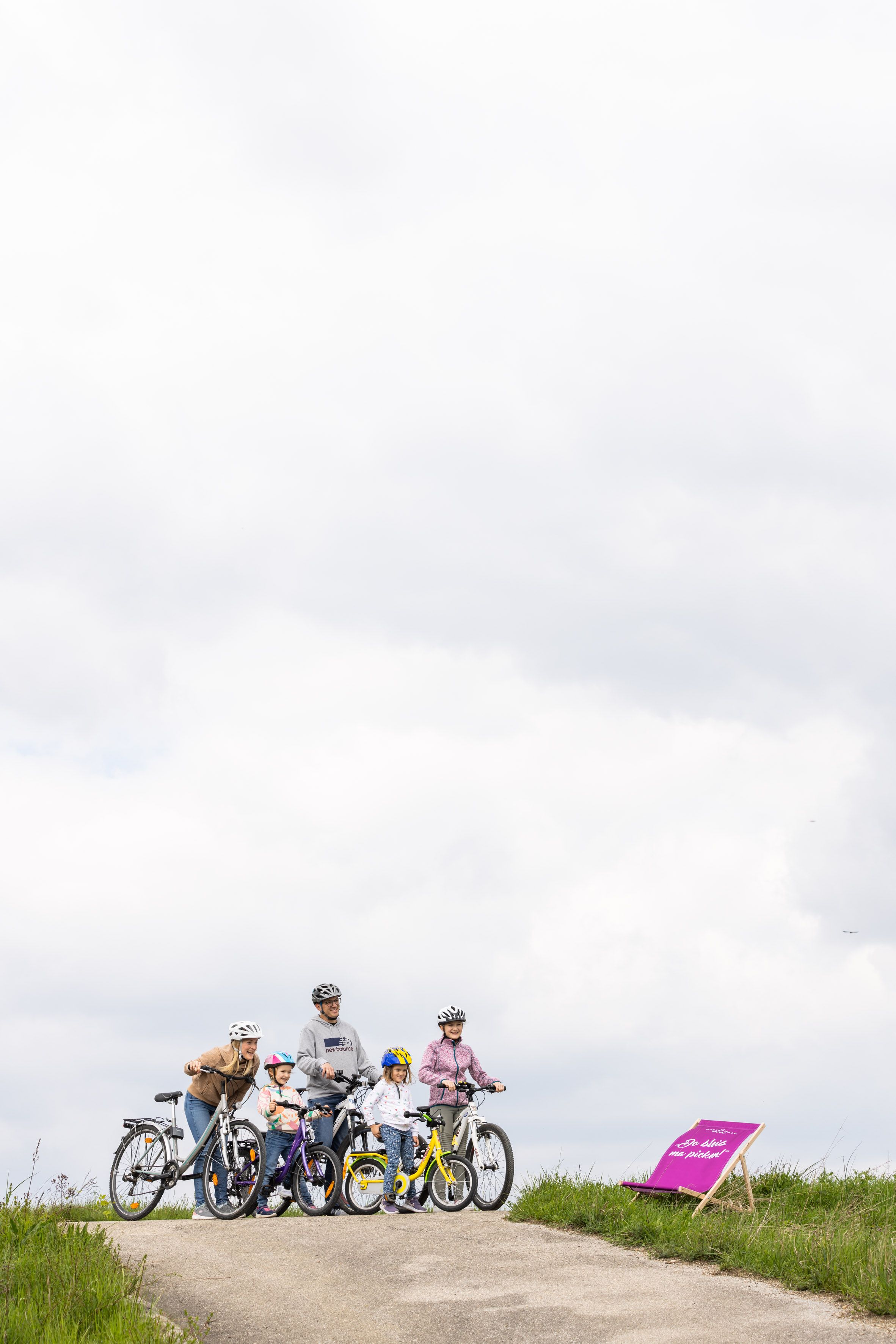 Eine fünfköpfige Familie, bestehend aus zwei Erwachsenen und drei Kindern, fährt mit Fahrrädern fröhlich auf einem Radweg, der sich entlang einer saftig grünen Wiese erstreckt.