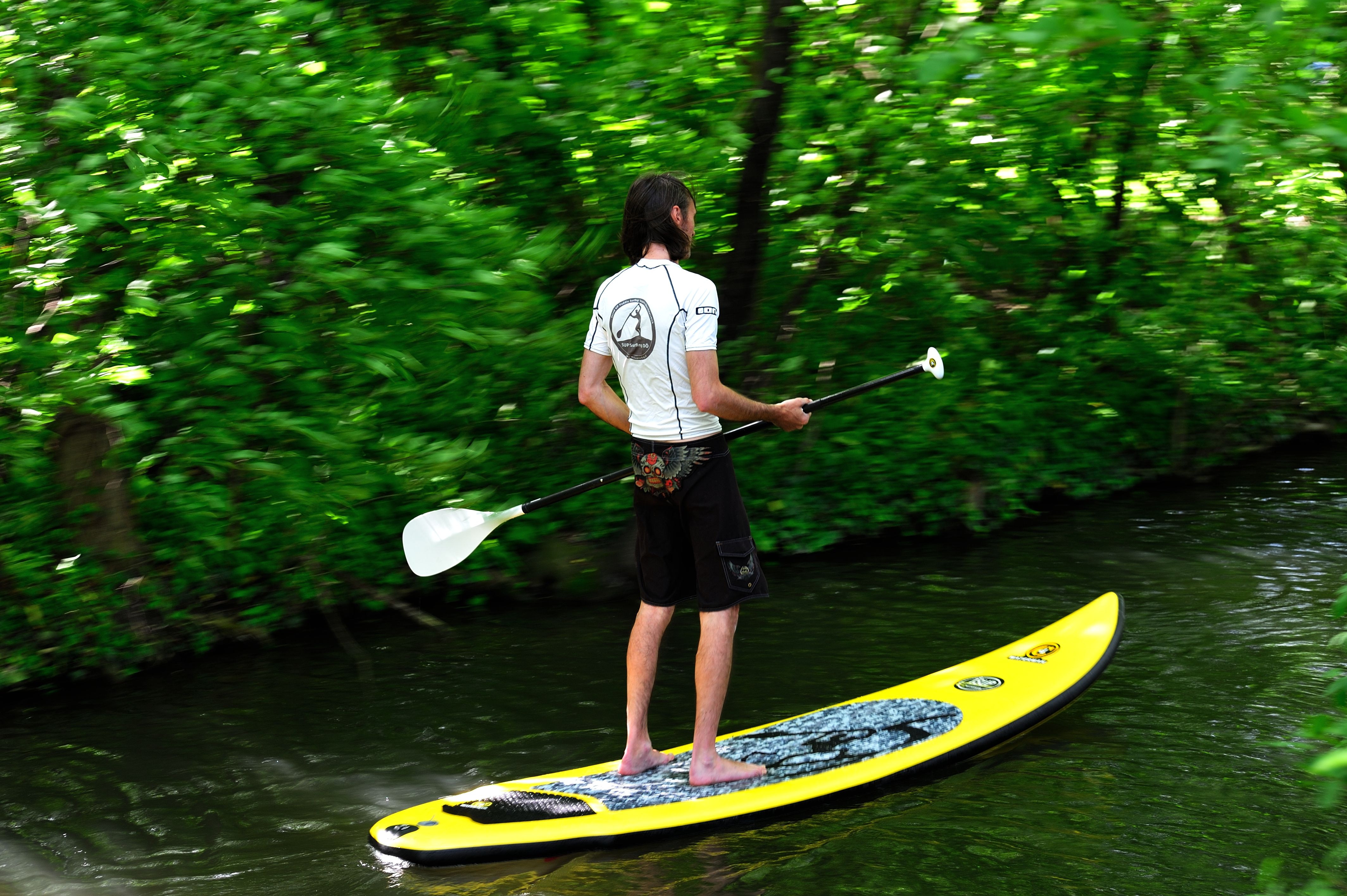 Person beim Stand Up Paddeln auf einem Fluss, umgeben von grüner Vegetation.