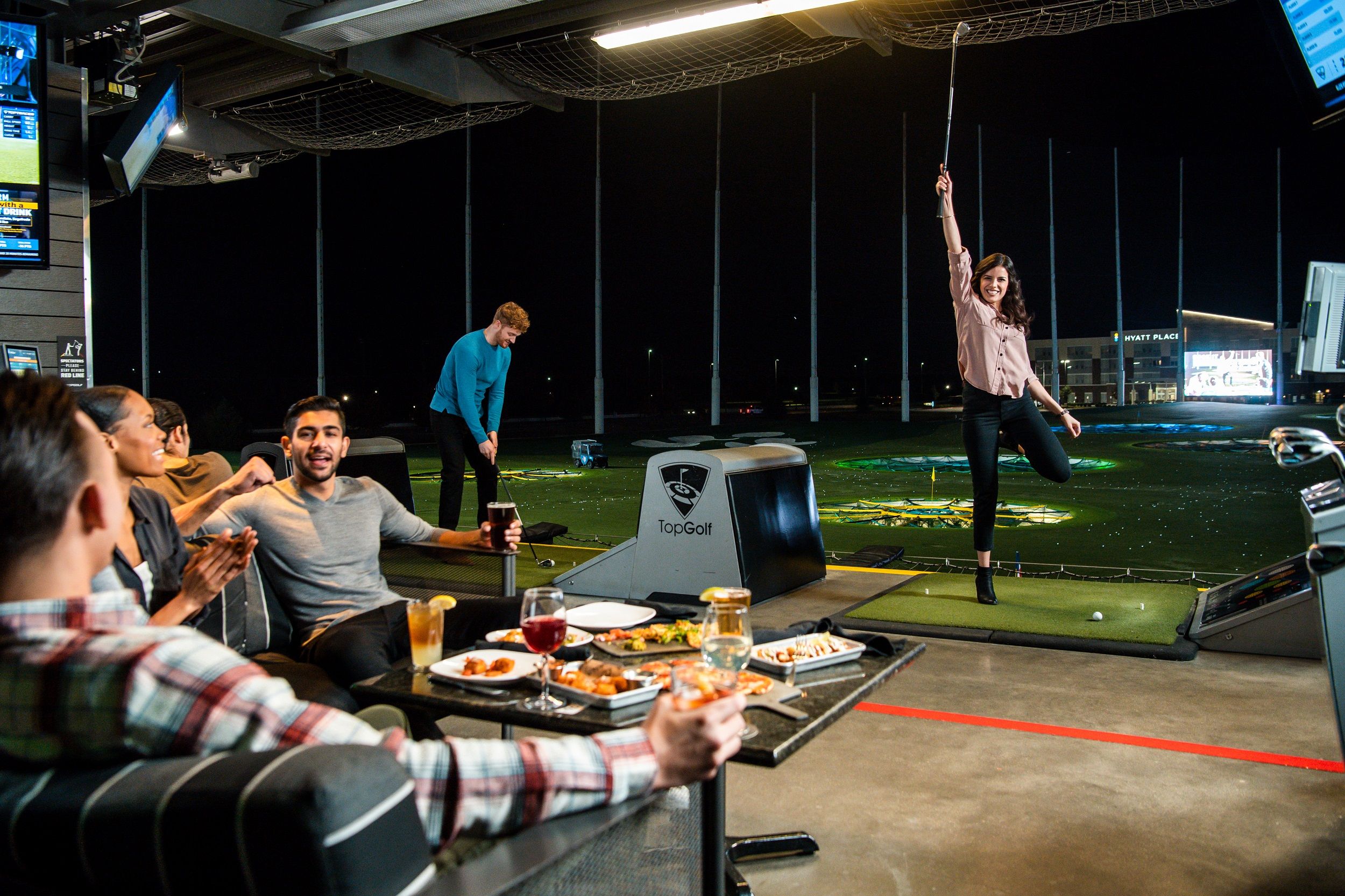 Menschen spielen Golf bei TopGolf, eine Frau jubelt, während andere essen und trinken.