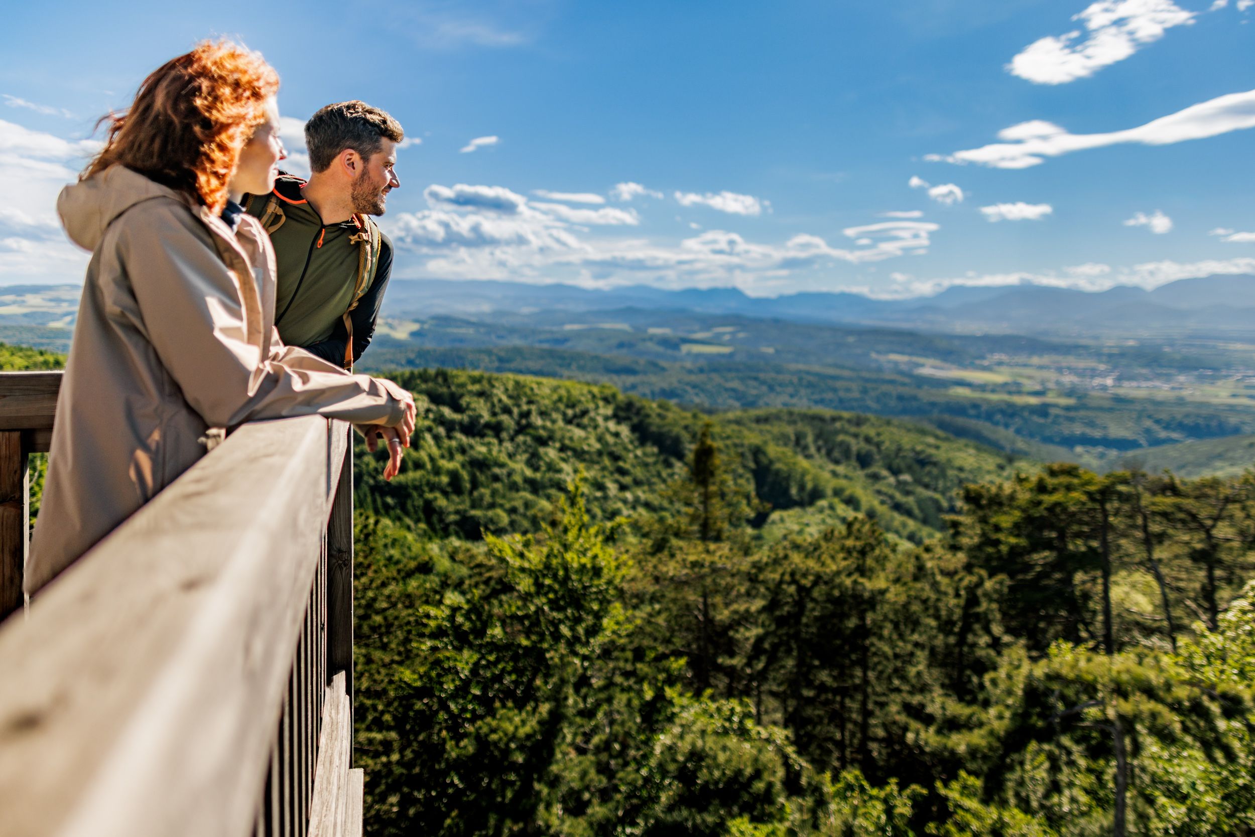 Zwei Personen stehen auf der Plattform des Aussichtsturmes Lanzenkirchen/Wiesen und blicken über eine bewaldete Landschaft mit Bergen im Hintergrund.