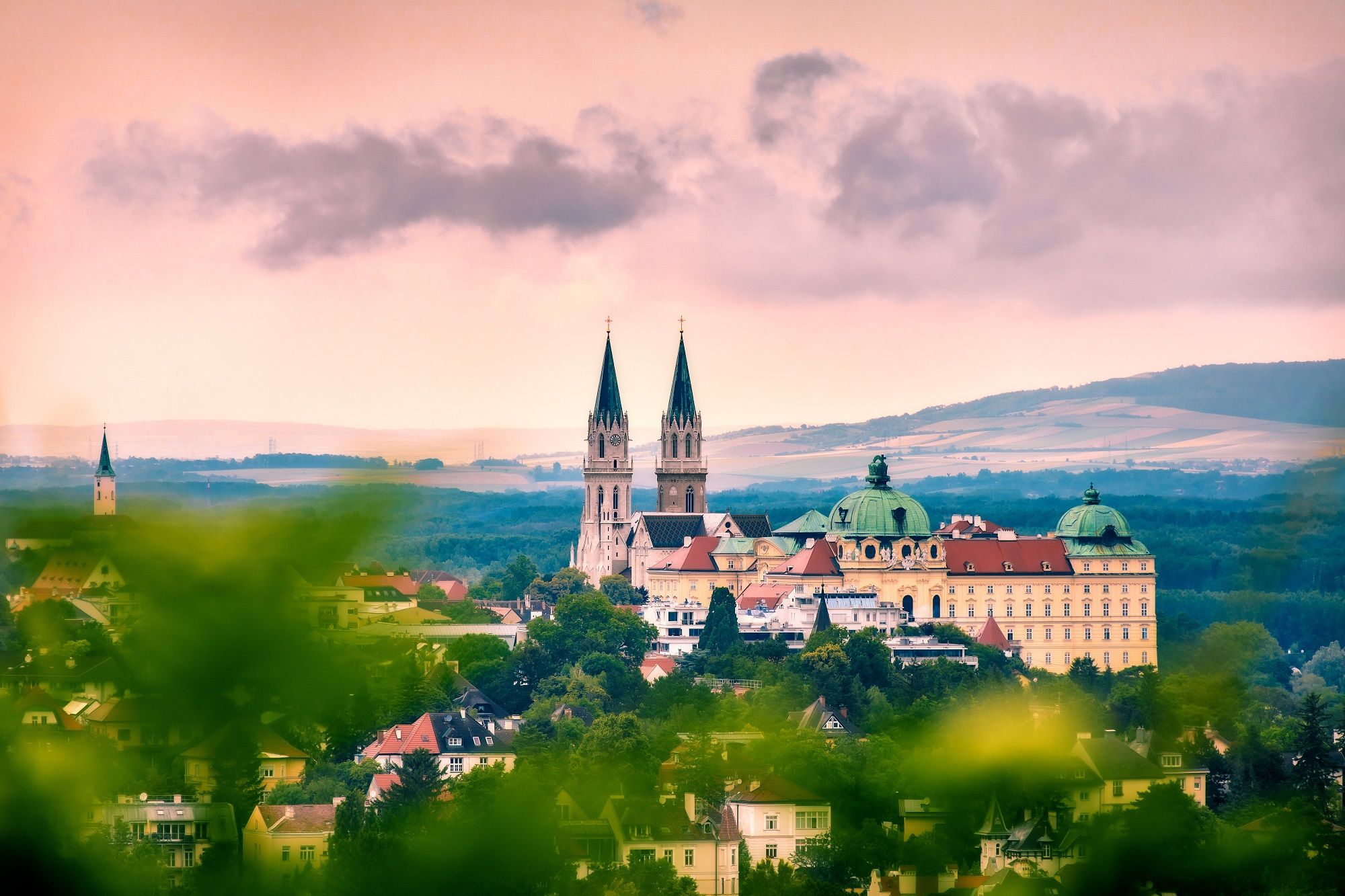 Panoramablick auf das Stift Klosterneuburg mit zwei Kirchtürmen und barockem Gebäude, umgeben von grüner Landschaft und unter bewölktem Himmel.