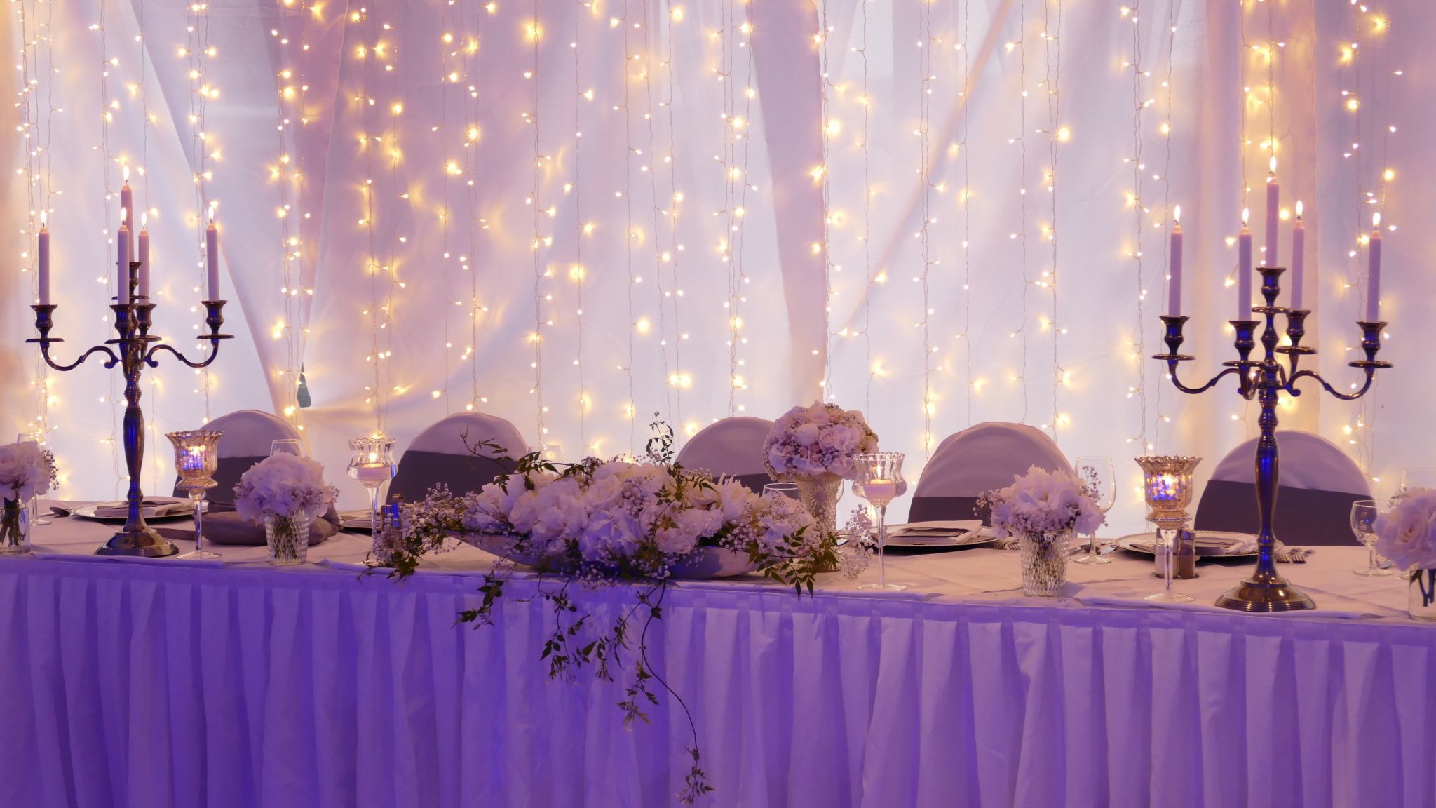 Elegante Tischdekoration mit Kerzenleuchtern und Blumen vor Lichterketten.