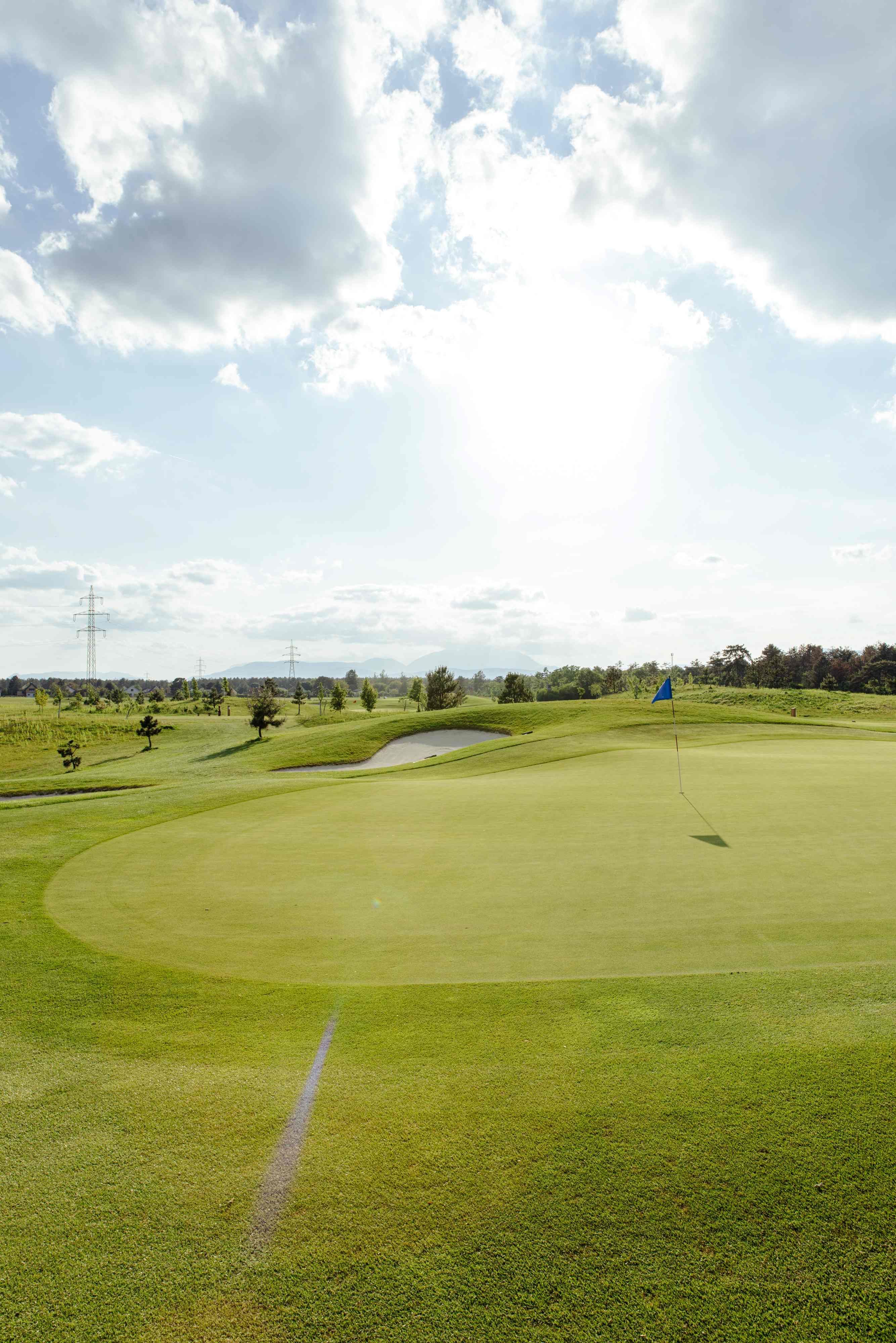 Golfplatz mit Fahne auf dem Grün, umgeben von Bäumen und Strommasten unter einem bewölkten Himmel.