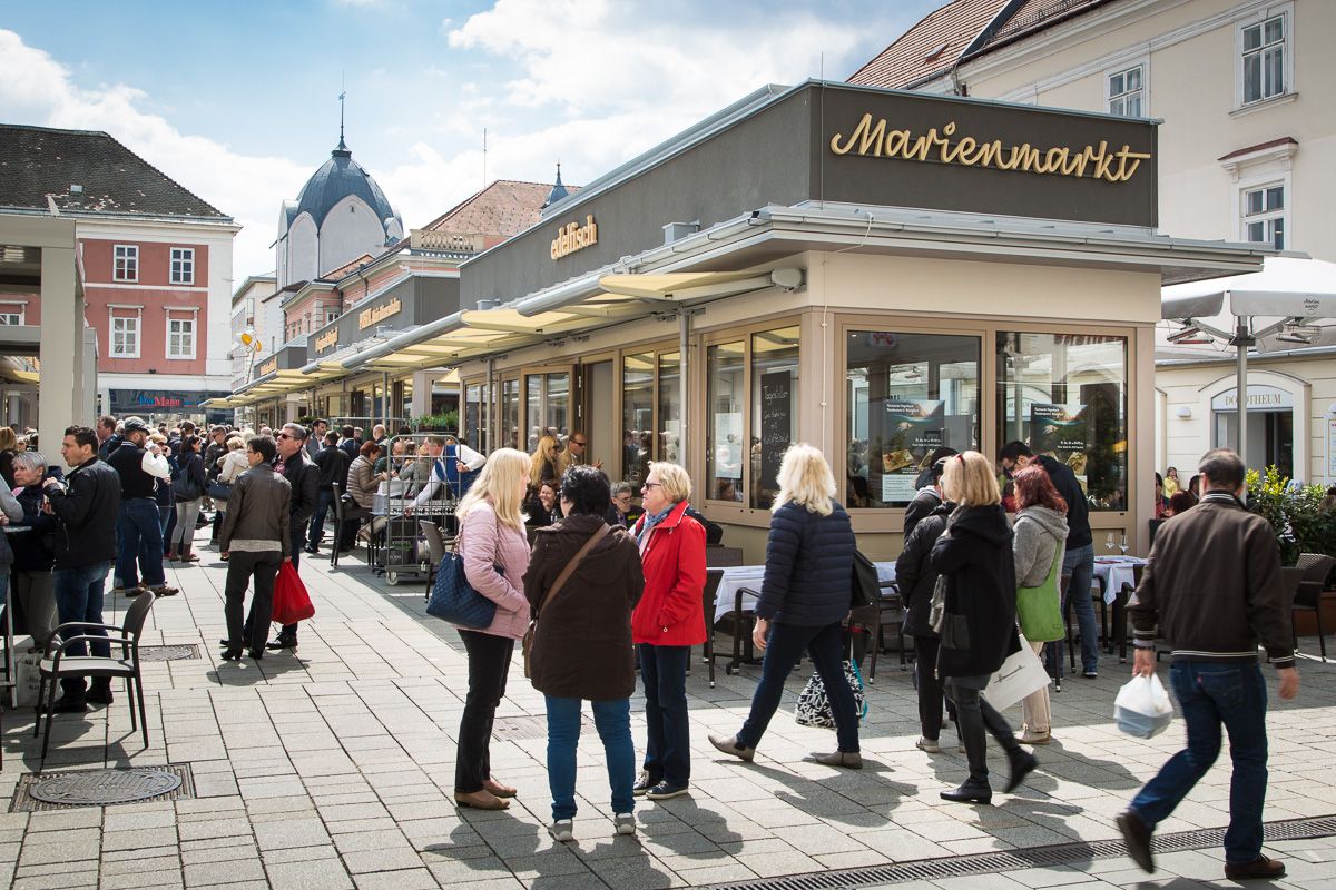 Menschenmenge auf einem belebten Markt mit Ständen und Gebäuden im Hintergrund.