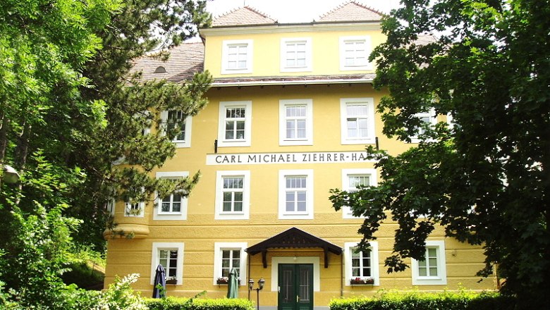 Frontansicht des Carl Michael Ziehrer-Hauses, ein gelbes Gebäude mit mehreren Fenstern und einem grünen Eingang, umgeben von Bäumen.