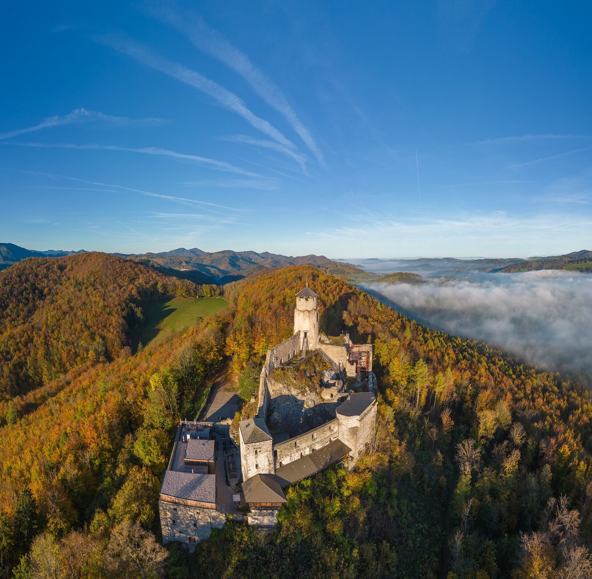 Luftaufnahme der Araburg im Herbst: Die Burg ist von orange- und gelb gefärbtem Wald umgeben, eine dichte Nebeldecke liegt über dem Tal, während oberhalb der Burg strahlender Sonnenschein den Himmel erhellt.