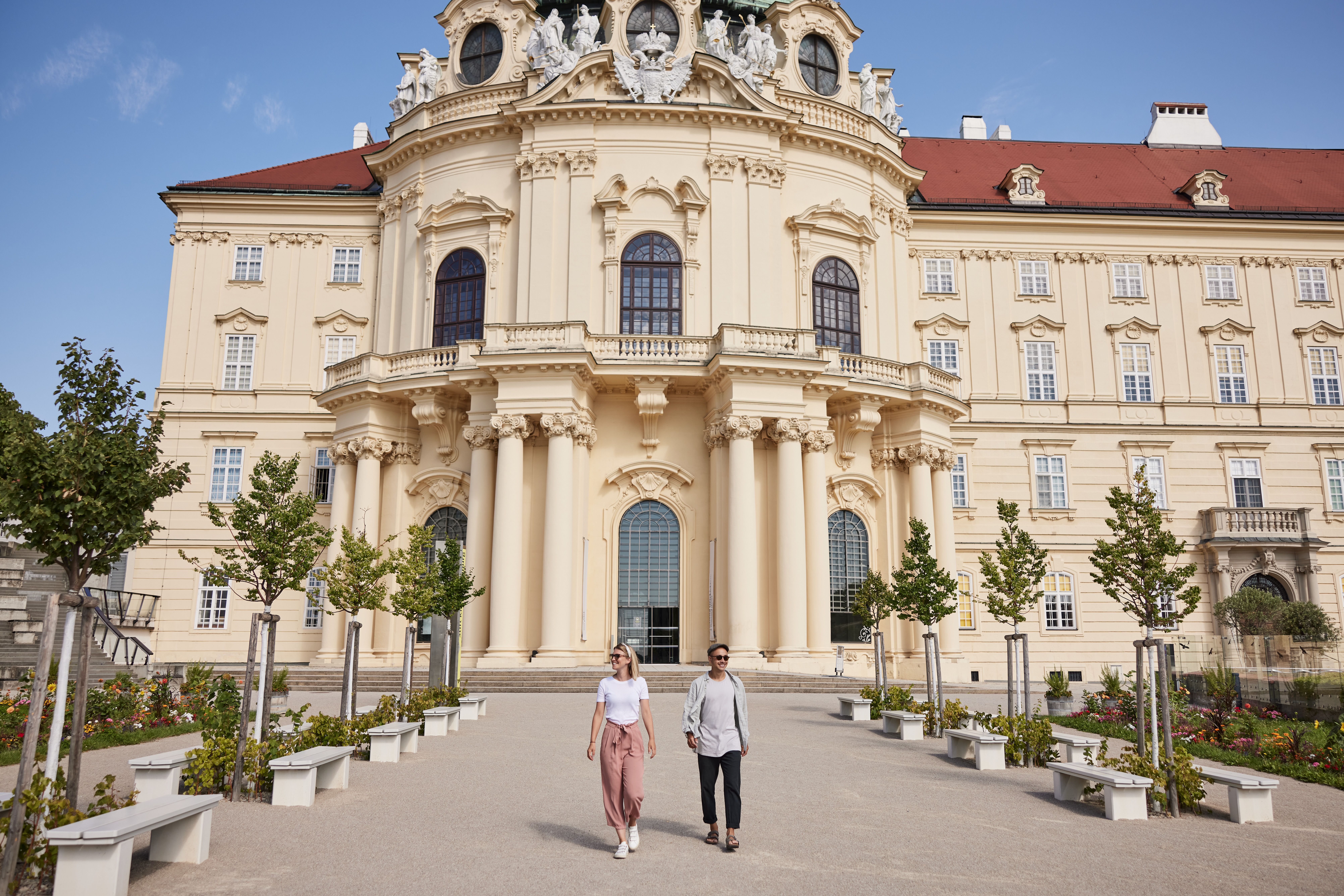 Ein sonniger Tag lädt zu einem entspannten Spaziergang ein, während die beeindruckende Architektur des Klosters im Hintergrund erstrahlt. Die blühenden Pflanzen und die gepflegte Umgebung schaffen eine harmonische Atmosphäre, die zum Verweilen einlädt.