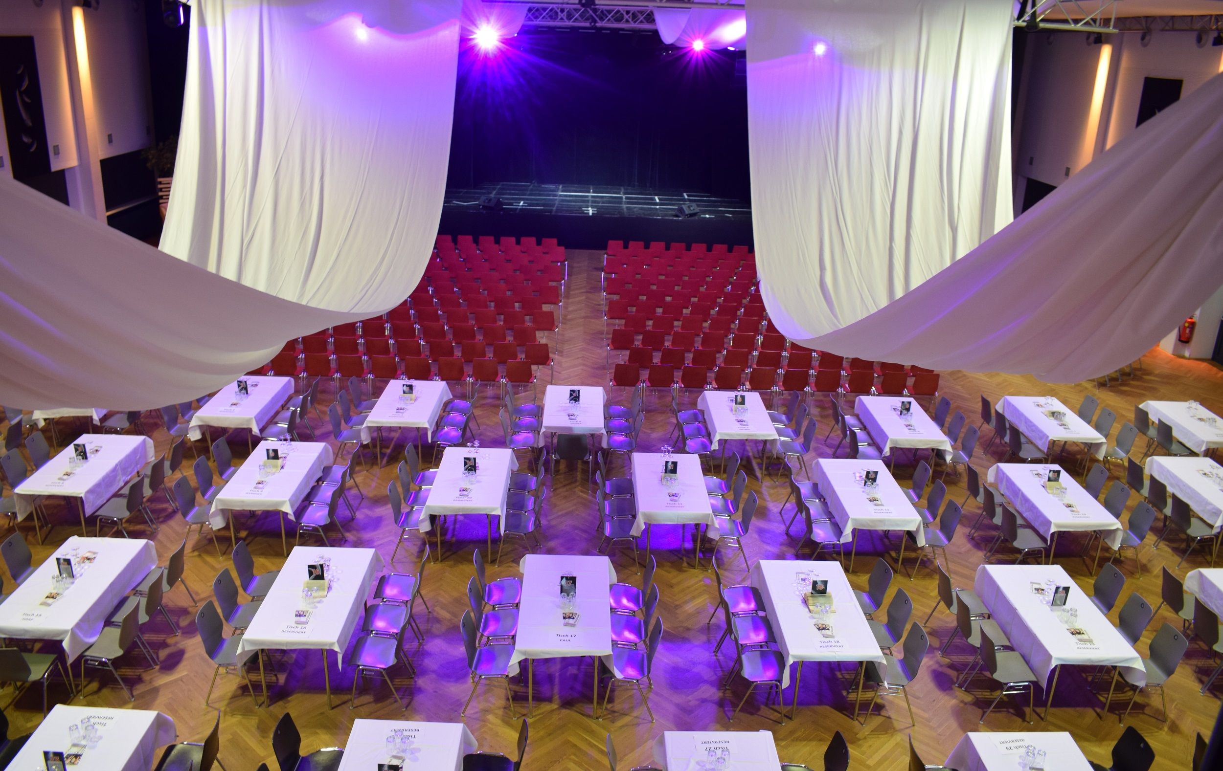 Ein Veranstaltungsraum mit Tischen und Stühlen, lila Beleuchtung und einer Bühne im Hintergrund.